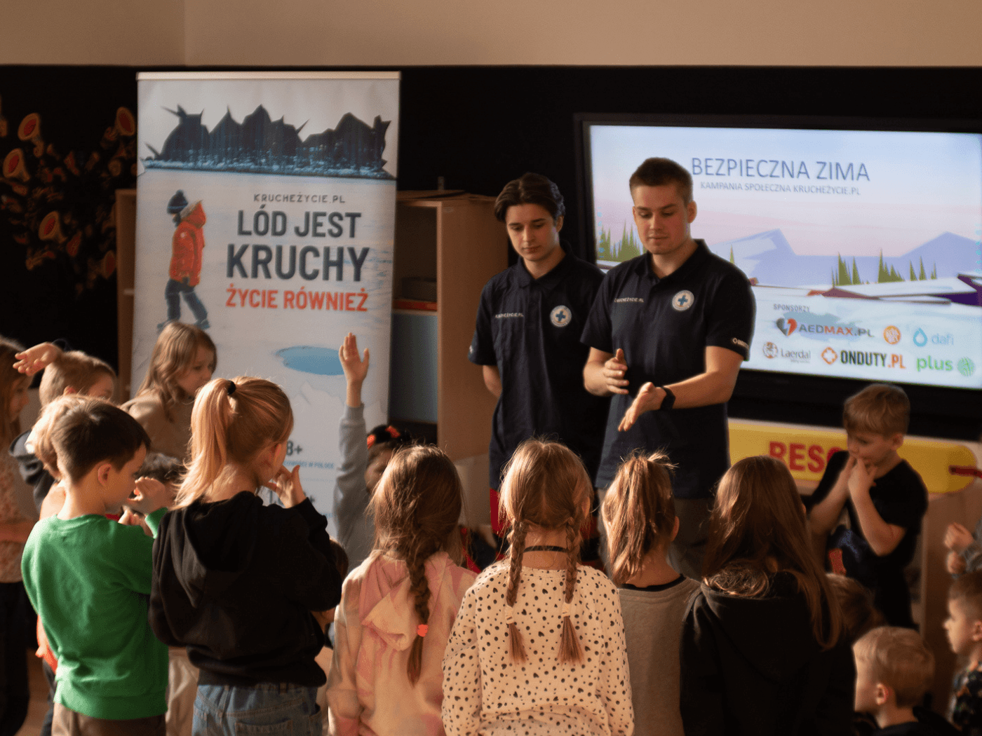 Dwóch ratowników WOPR prowadzi interaktywne zajęcia edukacyjne dla grupy dzieci w szkole podczas kampanii Kruche Życie – akcji zimowej promującej bezpieczeństwo na lodzie. W tle baner z hasłem „Lód jest kruchy, życie również".