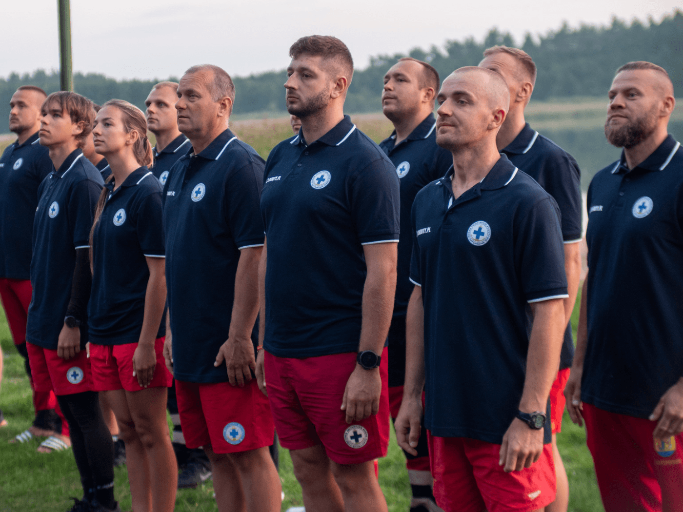 Grupa ratowników WOPR w granatowych koszulkach polo z logo organizacji i czerwonych szortach stojących w szeregu podczas uroczystości lub odprawy na świeżym powietrzu.