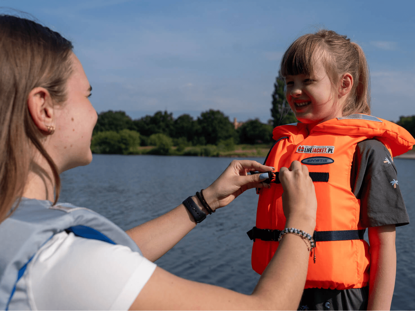 Wolontariuszka WOPR zapina pomarańczową kamizelkę ratunkową LifeJacket.pl na uśmiechniętej dziewczynce stojącej nad jeziorem podczas akcji edukacyjnej kampanii LifeJacket.
