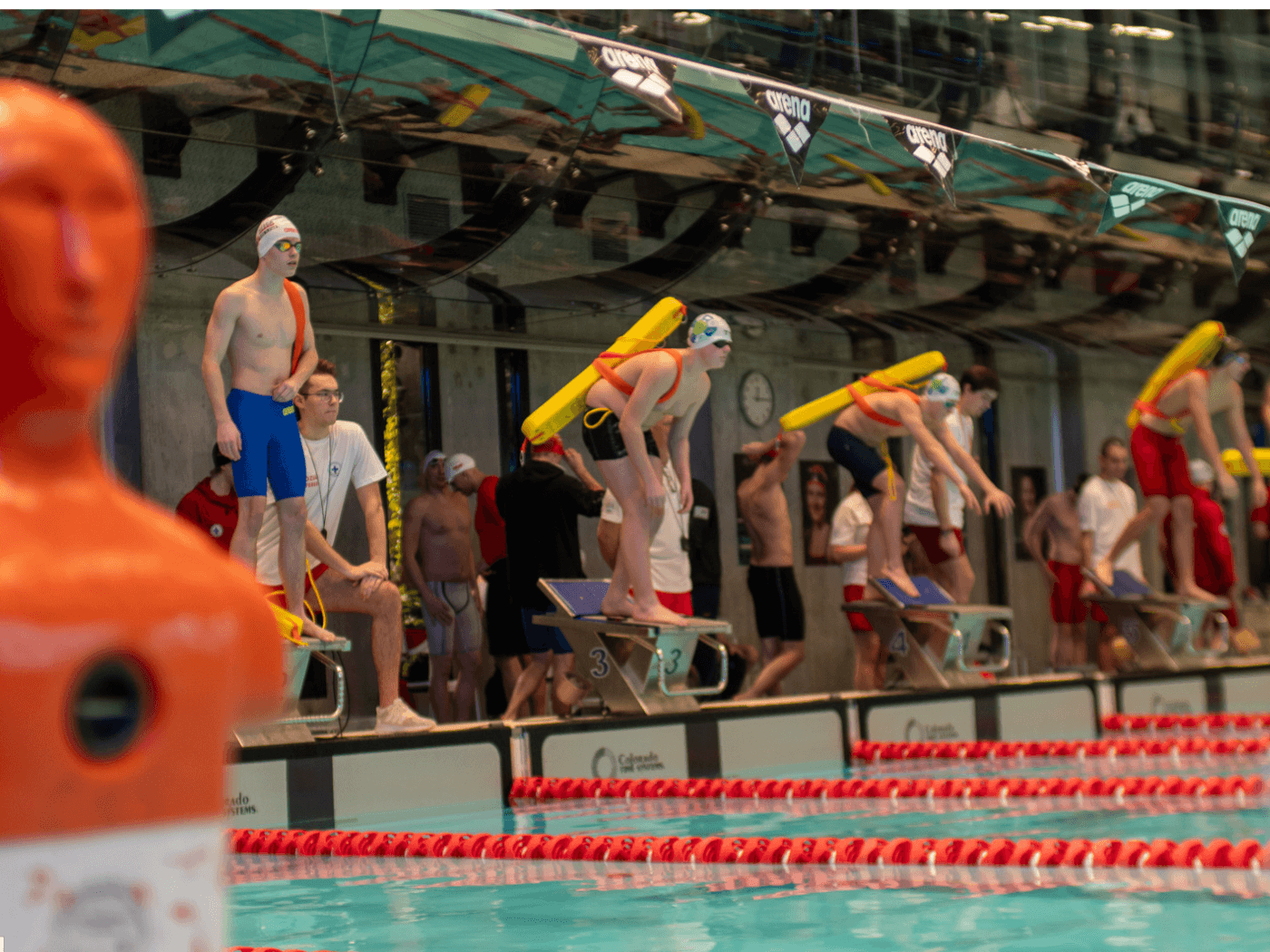 Zawodnicy sportowego ratownictwa wodnego stoją na blokach startowych z tubami ratunkowymi, gotowi do skoku na basenie podczas zawodów lifesaving organizowanych przez WOPR.